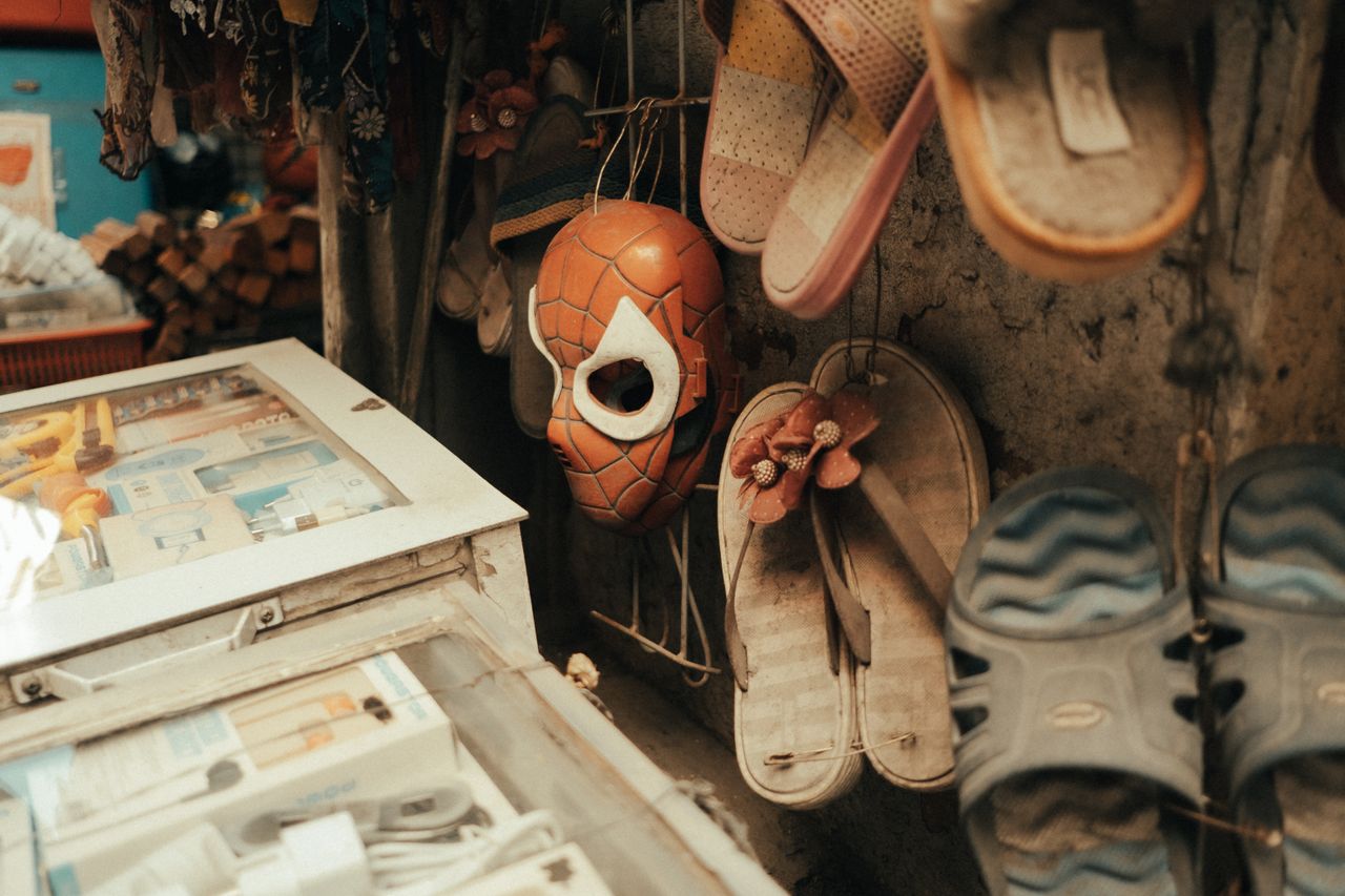 A rustic market stall displaying hanging flip flops and a Spiderman mask in a weathered setting.